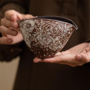 Könnte beinhalten: Ein brauner Keramik-Krug mit einem weißen Blumenmuster. Der Krug hat einen breiten, abgerundeten Boden und einen Ausguss auf einer Seite.