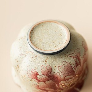 May include: A ceramic bowl with a floral design. The bowl is cream-colored with a red and gold peony pattern. The rim is a darker color, and the bowl has a textured surface.
