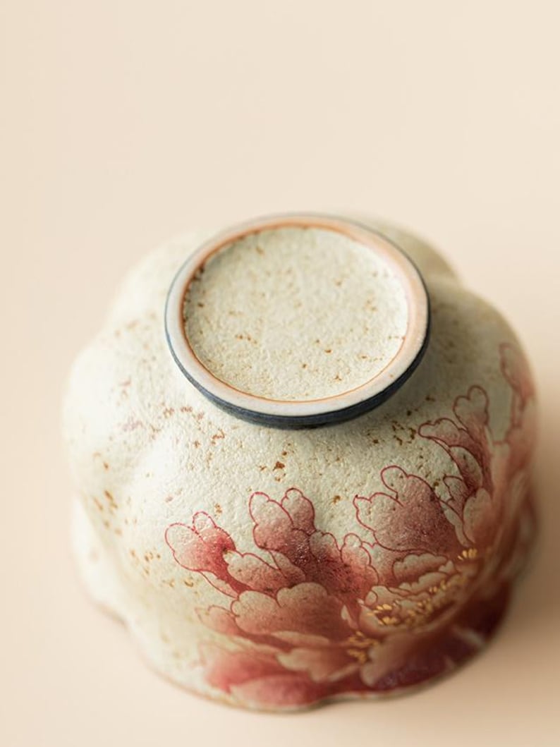 May include: A ceramic bowl with a floral design. The bowl is cream-colored with a red and gold peony pattern. The rim is a darker color, and the bowl has a textured surface.