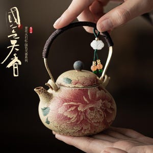 Peut inclure: Théière en céramique avec un motif floral rouge et crème. La théière a une anse marron foncé et un petit charme décoratif. Le texte sur le côté de la théière est dans une langue étrangère.