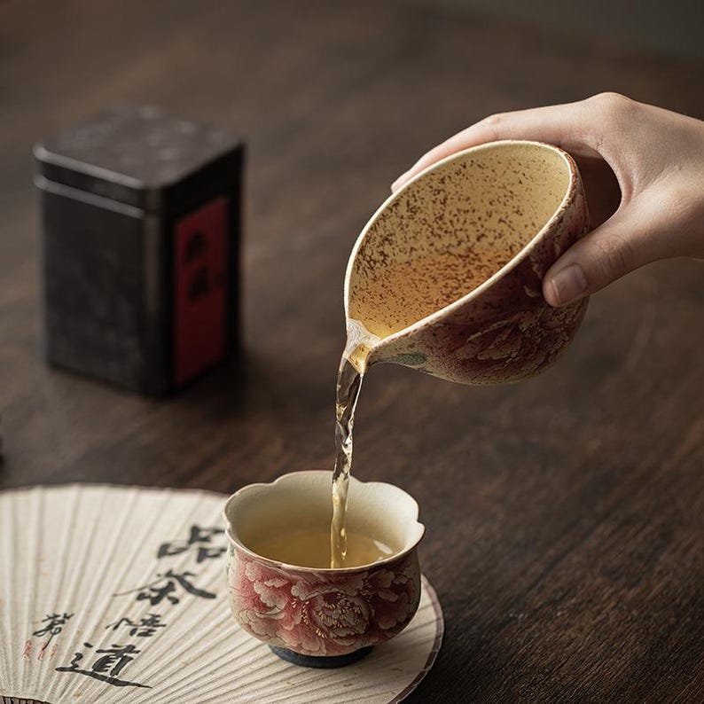May include: A hand pours tea from a ceramic pitcher into a matching cup. Both are decorated with red floral patterns. A black tea tin and a decorative fan with calligraphy are in the background. The tea is a light amber color.