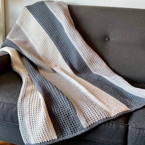 May include: A soft, textured blanket in shades of gray, white, and beige, draped over a dark gray sofa. The blanket features alternating stripes of solid and open-weave patterns, creating a cozy and inviting look. The blanket is approximately 60 inches wide and 70 inches long.