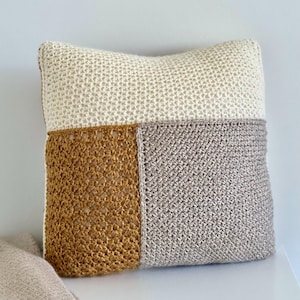 Può includere: Un cuscino quadrato decorativo con un design patchwork. La metà superiore è color crema, la parte inferiore sinistra è giallo senape e la parte inferiore destra è grigia. Il cuscino è realizzato in un materiale lavorato a uncinetto strutturato.