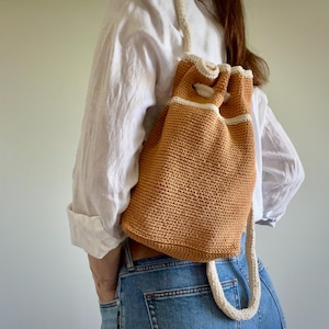 May include: A crocheted drawstring backpack in a warm orange hue, accented with cream-colored trim and a matching strap. The bag has a rounded shape and a textured knit pattern, offering a casual, handmade aesthetic. The bag is worn over the shoulder.