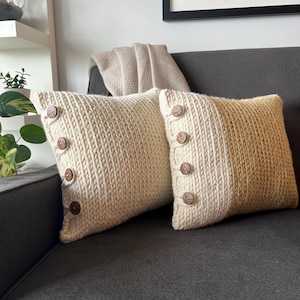 Puede incluir: Dos almohadas de punto con botones de madera. Una almohada es de color crema y la otra es crema con un degradado a un color marrón claro.