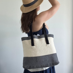 Puede incluir: Un bolso tote tejido con una sección central color crema y secciones superior e inferior grises. El bolso tiene asas negras y se lleva al hombro. Un accesorio elegante.