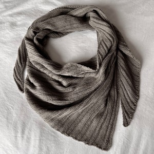 以下が含まれることがあります： 三角形の灰色のニットスカーフ。スカーフは折り畳まれて白い表面に置かれています