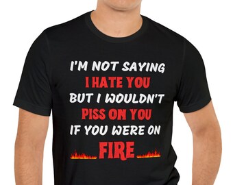 Op de afbeelding: Zwart T-shirt met een witte grafische print met de tekst "Ik zeg niet dat ik je haat, maar ik zou niet op je plassen als je in brand stond". De tekst is rood en heeft een vlammengrafiek eronder.