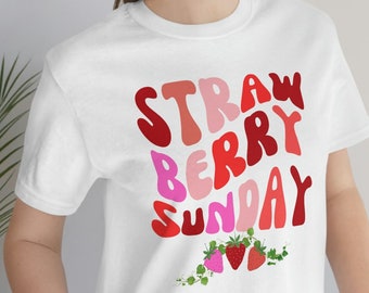 Op de afbeelding: Wit T-shirt met een grafisch ontwerp dat "Strawberry Sunday" zegt in een retro lettertype met rode, roze en witte kleuren. De grafiek bevat ook een kleine groep rode aardbeien met groene bladeren.