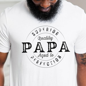 Peut inclure: T-shirt blanc avec un motif noir circulaire indiquant "SUPERIOR Quality PAPA Aged to PERFECTION". Le t-shirt est de style col rond.