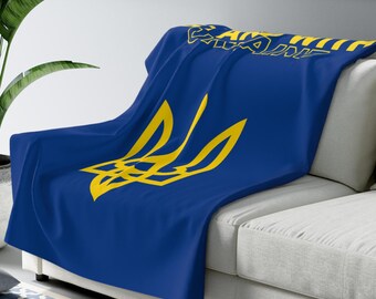 May include: A blue blanket with a yellow trident symbol and the word "UKRAINE" in yellow lettering. The blanket is draped over a white sofa. A large green plant is in the background.