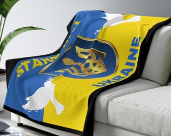 May include: A blue and yellow blanket with the words "STAND WITH UKRAINE" in gold lettering. The blanket features a gold emblem and a white dove design. The blanket is draped over a white couch.