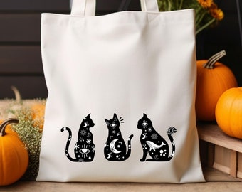May include: White canvas tote bag with three black silhouette cats with celestial designs. The cats are sitting in a row. The bag is on a wooden surface with pumpkins in the background.