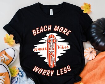 May include: Black t-shirt featuring a graphic of a skeleton surfing on a surfboard. The text "Beach More Summer Vibes Worry Less" is printed on the shirt.