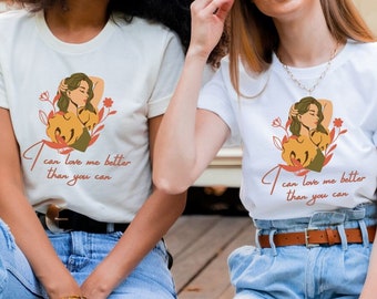 Könnte beinhalten: Zwei Personen tragen weiße T-Shirts mit einem Grafikdruck einer Frau und dem Text "Ich kann mich besser lieben als du". Sie sitzen auf einem Holzdeck vor einem Wohnmobil.