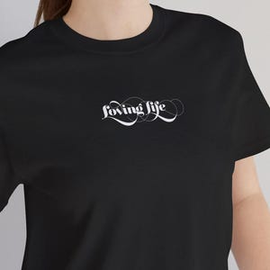 May include: A black short-sleeved t-shirt featuring the white text "loving life" in a cursive, swirling script font centered on the chest. The garment has a classic crew neckline and a straight hem, providing a simple and clean aesthetic.