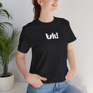 May include: Black t-shirt with the white text "tsk!". The shirt has short sleeves and a crew neck. The model is wearing blue jeans. The shirt is made of a soft, comfortable material.