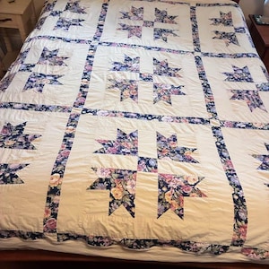 May include: A cream-colored quilt with a star pattern. The stars are filled with floral fabric in shades of pink, purple, and yellow. The quilt has a dark blue floral border and is on a wooden bed frame.