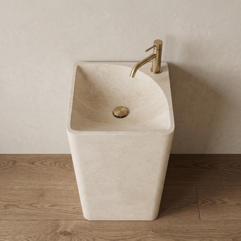 May include: A modern, freestanding bathroom sink made of light beige stone. It features a square basin with a curved edge, a gold-colored faucet, and a matching drain cover. The sink is set against a neutral wall and wooden floor.