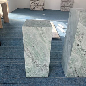 Green marble pedestal pair displayed in a minimalist room with natural light. Their tall and short shapes are emphasized by unique veining and smooth finishes. These plinths offer functional elegance, suitable for showcasing floral arrangements, busts, or art pieces. Perfect for design-focused homes, creative studios, or gallery spaces. Crafted from real marble, these stands highlight the natural beauty of stone in a bold and modern silhouette.
