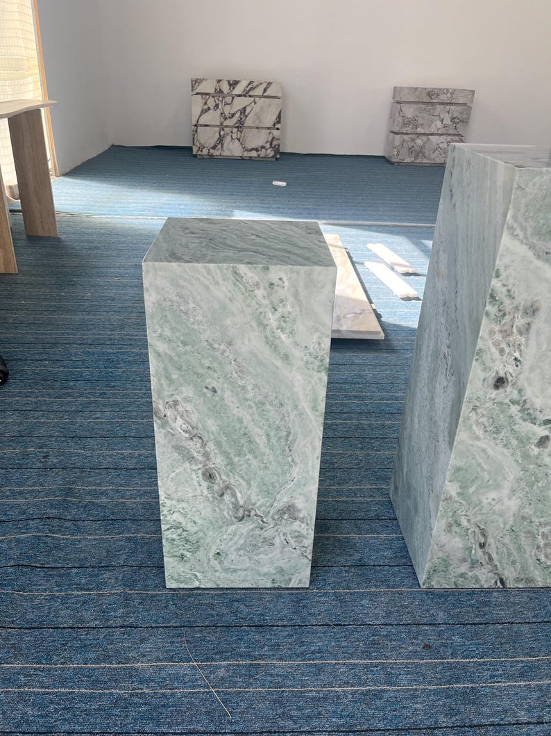 Green marble pedestal pair displayed in a minimalist room with natural light. Their tall and short shapes are emphasized by unique veining and smooth finishes. These plinths offer functional elegance, suitable for showcasing floral arrangements, busts, or art pieces. Perfect for design-focused homes, creative studios, or gallery spaces. Crafted from real marble, these stands highlight the natural beauty of stone in a bold and modern silhouette.