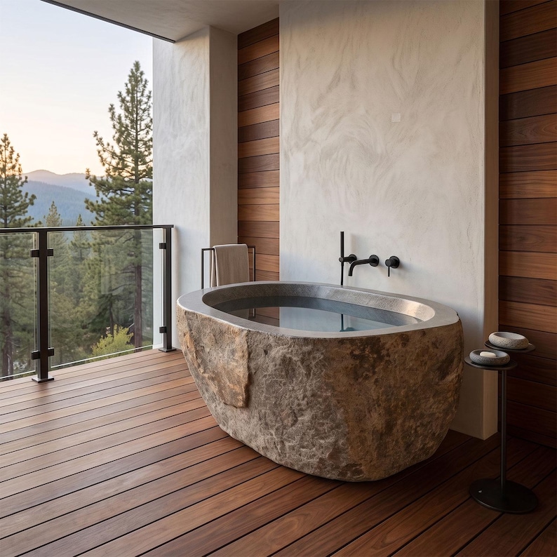 May include: An oval-shaped bathtub carved from a single piece of natural stone with a rough, textured exterior and a smooth, polished interior basin. The tub features matte black wall-mounted plumbing fixtures and is positioned on a wooden deck.