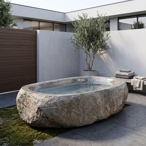 May include: An oval-shaped bathtub carved from a single piece of natural beige stone with a rough, textured exterior and a smooth, polished interior basin filled with clear water, positioned outdoors on a paved surface near a small tree.