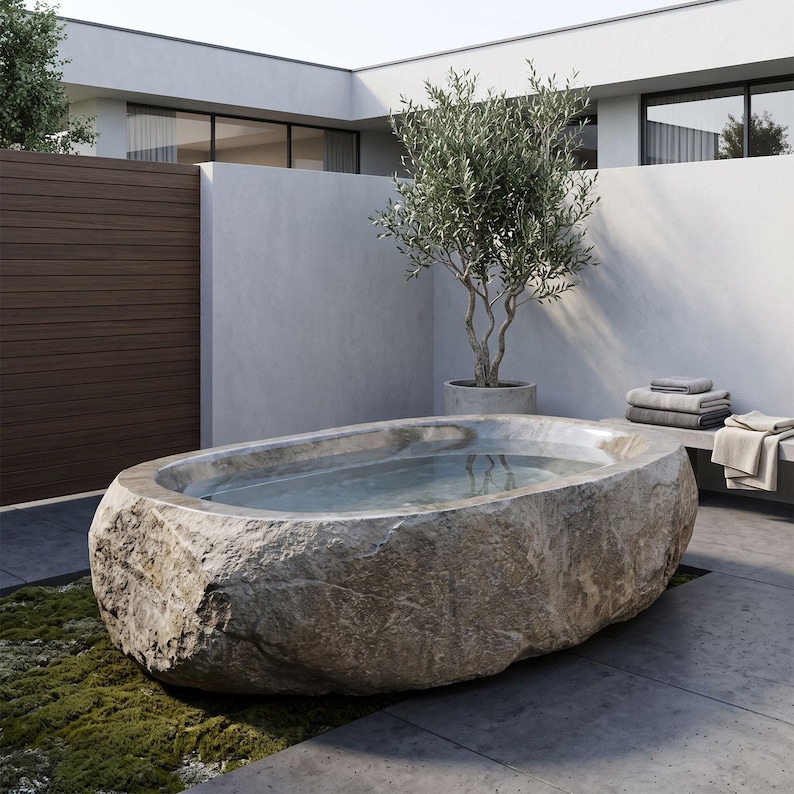 May include: An oval-shaped bathtub carved from a single piece of natural beige stone with a rough, textured exterior and a smooth, polished interior basin filled with clear water, positioned outdoors on a paved surface near a small tree.