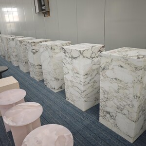 May include: A row of white marble pedestals of varying heights, arranged in a room with a light blue carpet. The pedestals are made of white marble with gray veining.