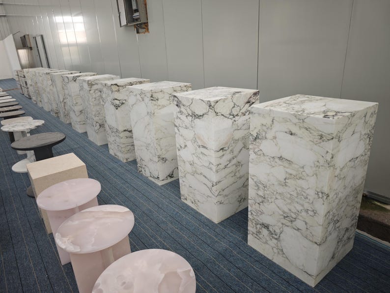 May include: A row of white marble pedestals of varying heights, arranged in a room with a light blue carpet. The pedestals are made of white marble with gray veining.