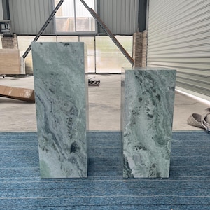 Green marble pedestal pair displayed in a minimalist room with natural light. Their tall and short shapes are emphasized by unique veining and smooth finishes. These plinths offer functional elegance, suitable for showcasing floral arrangements, busts, or art pieces. Perfect for design-focused homes, creative studios, or gallery spaces. Crafted from real marble, these stands highlight the natural beauty of stone in a bold and modern silhouette.