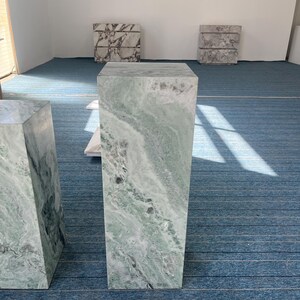 Front view of a short green marble pedestal featuring flowing light and dark veining. The stone has a soft matte finish and natural texture, ideal for modern or vintage-style interiors. Use it as a decorative side table, display column, or plant stand. Its sculptural form adds visual interest to any corner or entryway. Made from solid stone, this durable piece merges classic natural materials with modern furniture design for timeless style.