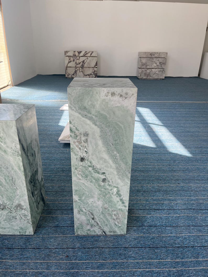 Front view of a short green marble pedestal featuring flowing light and dark veining. The stone has a soft matte finish and natural texture, ideal for modern or vintage-style interiors. Use it as a decorative side table, display column, or plant stand. Its sculptural form adds visual interest to any corner or entryway. Made from solid stone, this durable piece merges classic natural materials with modern furniture design for timeless style.