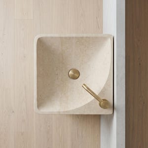 May include: A square, beige stone sink with rounded corners. It features a circular, gold-colored drain cover and a matching gold faucet. The sink is set against a light wood floor and a white wall.