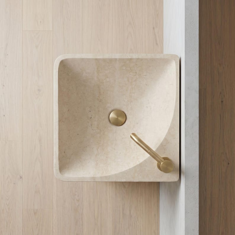 May include: A square, beige stone sink with rounded corners. It features a circular, gold-colored drain cover and a matching gold faucet. The sink is set against a light wood floor and a white wall.