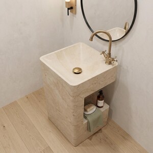 May include: A beige stone pedestal sink with a gold faucet and drain. A small shelf holds a soap dish, a green towel, and a brown bottle. The sink is set against a light-colored wall with a round mirror.