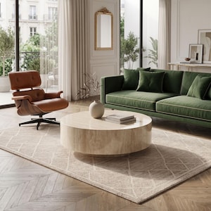 May include: A round, beige travertine coffee table with a smooth, polished surface sits on a textured area rug. The table features a cylindrical base and a flat top, holding a small ceramic vase with branches and two stacked books.