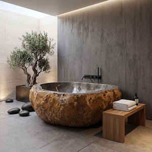 May include: A freestanding bathtub carved from a large, textured brown stone with a smooth, polished metallic interior basin. The tub is positioned in a room with gray concrete walls, a wooden bench with white towels, and a potted olive tree.