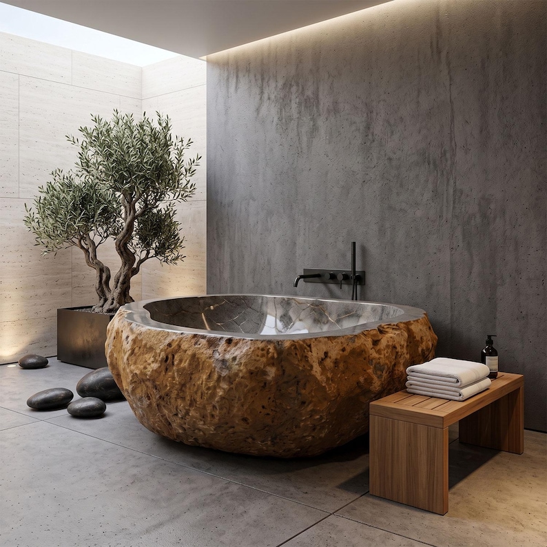 May include: A freestanding bathtub carved from a large, textured brown stone with a smooth, polished metallic interior basin. The tub is positioned in a room with gray concrete walls, a wooden bench with white towels, and a potted olive tree.