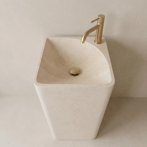 May include: A modern, cream-colored, rectangular sink with a curved top and a gold faucet. The sink has a gold drain stopper. The faucet is positioned on the right side of the sink.