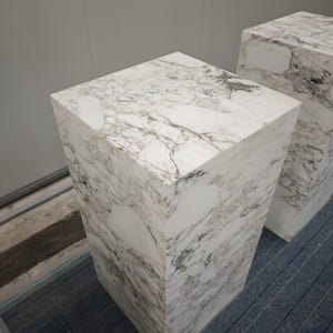 May include: Two white marble pedestals with a veined pattern. The pedestals are square and have a smooth, polished surface.