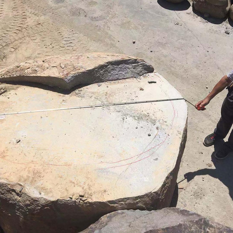 May include: A large, light beige, irregularly shaped stone slab with a rough, textured surface. A white measuring tape is stretched across the top of the stone, showing a measurement of approximately 60 inches from edge to edge.