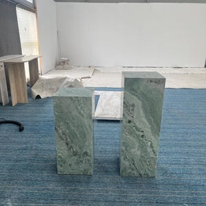 Pair of green onyx marble pedestal stands shown on a blue carpet in a studio setting. These tall rectangular plinths feature dramatic natural veining in shades of mint, gray, and white. Ideal for use as sculpture pedestals, plant stands, or entryway columns. The polished stone finish enhances their luxurious feel, making them perfect for high-end interiors. These solid marble pieces blend vintage elegance with modern design aesthetics.