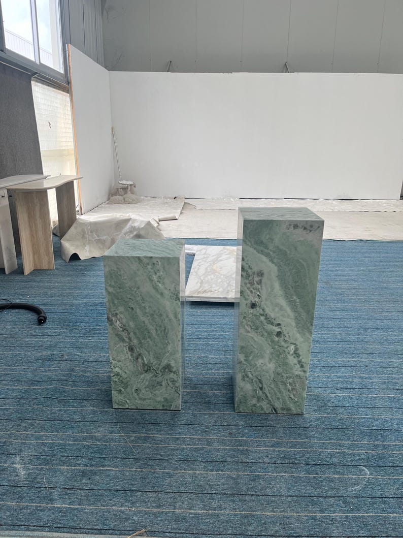Pair of green onyx marble pedestal stands shown on a blue carpet in a studio setting. These tall rectangular plinths feature dramatic natural veining in shades of mint, gray, and white. Ideal for use as sculpture pedestals, plant stands, or entryway columns. The polished stone finish enhances their luxurious feel, making them perfect for high-end interiors. These solid marble pieces blend vintage elegance with modern design aesthetics.