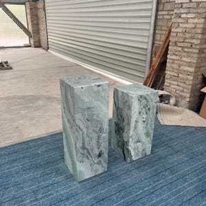 Angled view of two polished green onyx marble pedestal stands with blue carpet beneath, showing off their elegant proportions and natural veining. The taller and shorter plinths have complementary textures, adding layered visual interest. Their minimalist geometric form works well in contemporary spaces, luxury boutiques, or home interiors as display stands, side tables, or modern sculpture bases. Durable and stylish, these pedestals elevate any room.