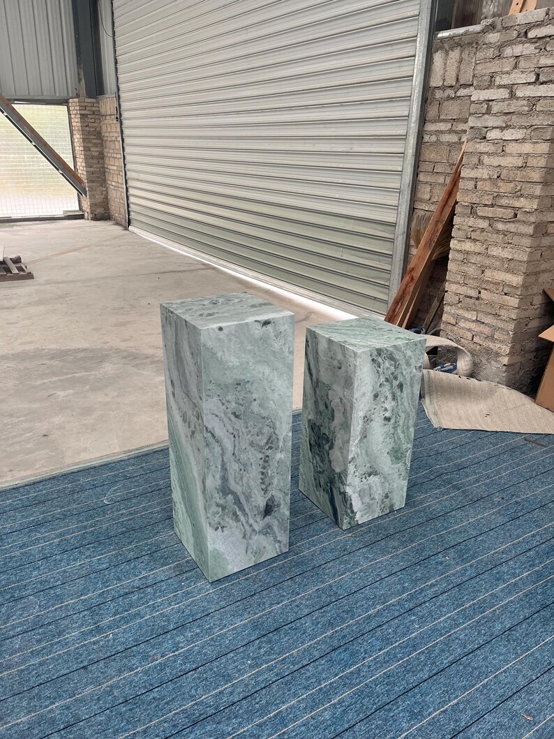 Angled view of two polished green onyx marble pedestal stands with blue carpet beneath, showing off their elegant proportions and natural veining. The taller and shorter plinths have complementary textures, adding layered visual interest. Their minimalist geometric form works well in contemporary spaces, luxury boutiques, or home interiors as display stands, side tables, or modern sculpture bases. Durable and stylish, these pedestals elevate any room.