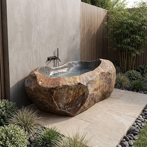 May include: A freestanding bathtub carved from a large, natural brown and tan boulder with a smooth, polished interior basin. The tub is positioned against a smooth gray concrete wall with a dark metal wall-mounted faucet fixture above it.