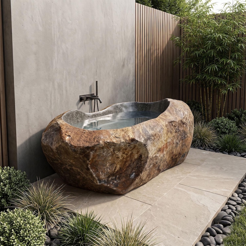 May include: A freestanding bathtub carved from a large, natural brown and tan boulder with a smooth, polished interior basin. The tub is positioned against a smooth gray concrete wall with a dark metal wall-mounted faucet fixture above it.