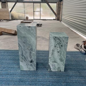 Two green marble pedestal tables, one tall and one shorter, placed in an industrial-style setting with large windows. Each column features swirling natural patterns unique to onyx marble, making every piece one of a kind. Their sculptural silhouette and durable stone material make them ideal for displaying art, flowers, or collectibles. Add luxury to any space with these handcrafted pedestals, perfect for entryways, studios, or modern gallery-inspired interiors.