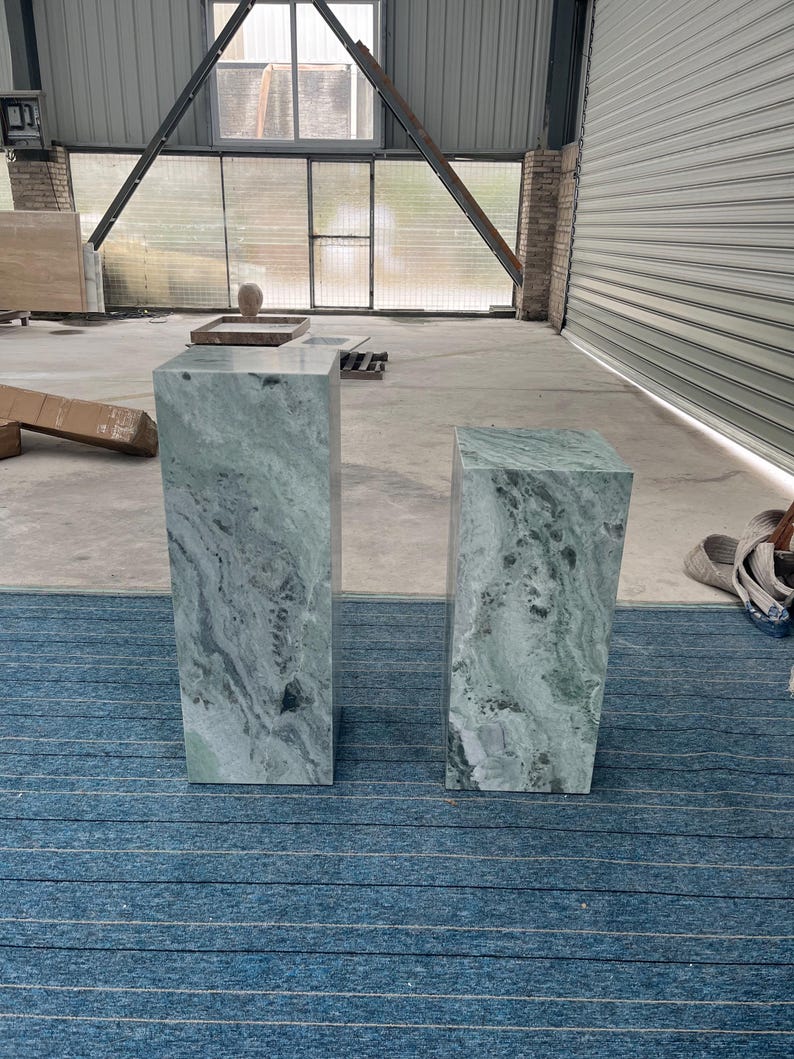 Two green marble pedestal tables, one tall and one shorter, placed in an industrial-style setting with large windows. Each column features swirling natural patterns unique to onyx marble, making every piece one of a kind. Their sculptural silhouette and durable stone material make them ideal for displaying art, flowers, or collectibles. Add luxury to any space with these handcrafted pedestals, perfect for entryways, studios, or modern gallery-inspired interiors.